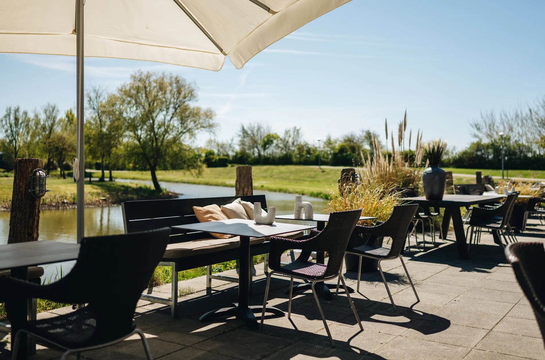 Sfeervol buitenterras aan het water met loungebanken, donkere stoelen en een weids uitzicht op het groene landschap onder een strakblauwe lucht.